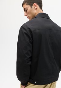 Schwarze Jacke aus glattem Stoff mit klassischem Kragen. Verfügt über ein schlichtes, gerades Design und gerippte Bündchen. Dezente Hardware-Detaillierung am Saum.