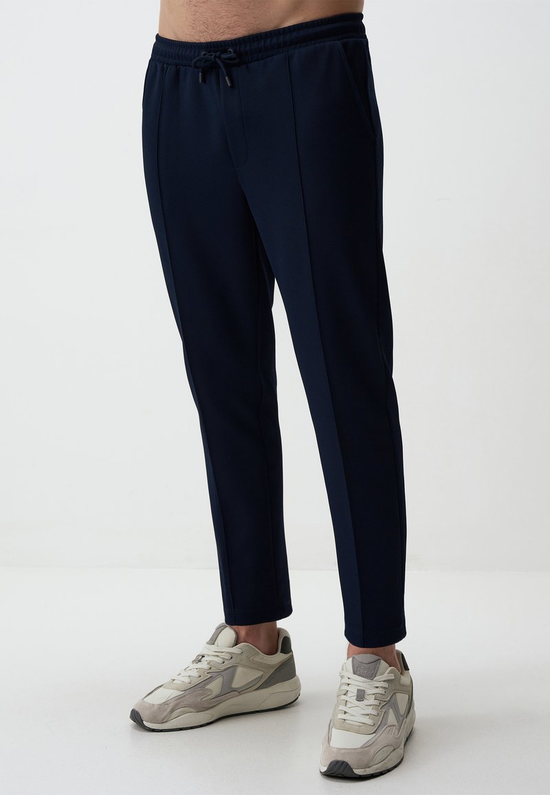 Pantalons de sport navy avec une taille à cordon de serrage, coupe ajustée, poches latérales et texture lisse. Associés à des baskets grises et blanches.