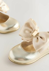 Ballerines métalliques dorées pour enfants avec grands nœuds en satin beige et une petite étiquette dorée sur un nœud portant l'inscription « BAKER ».