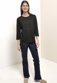 Schwarzes Langarmshirt mit grafischem Text, kombiniert mit dunkelblauen Jeans. Das Outfit wird durch hellgraue Sneaker ergänzt, vor einem neutralen Hintergrund.