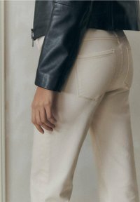 Jeans beige con dettagli di cucitura sottili, abbinati a una giacca di pelle nera attillata. Focus sulla vita e sulle tasche posteriori visibili.