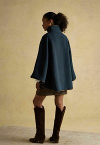 Žena s kučeravými vlasmi v cope, oblečená v tmavomodrom plášti, károvaných sukni a vysokých hnedých semišových čižmách, stojaca pred béžovým pozadím.