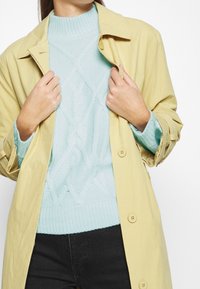 Gul trenchcoat med krage, bärs över en ljusblå kabelstickad tröja. Mörka byxor synliga nedtill. Texturerade material är tydliga.