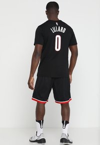 Svart baskettröja med "LILLARD" och nummer "0" på baksidan, i kombination med svarta shorts med röda detaljer och vita sneakers.