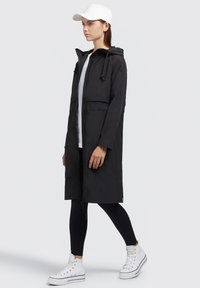 Longue veste noire avec capuche, fermeture éclair et poches avant. Associée à des leggings noirs et à des baskets montantes blanches, complétée par une casquette blanche.