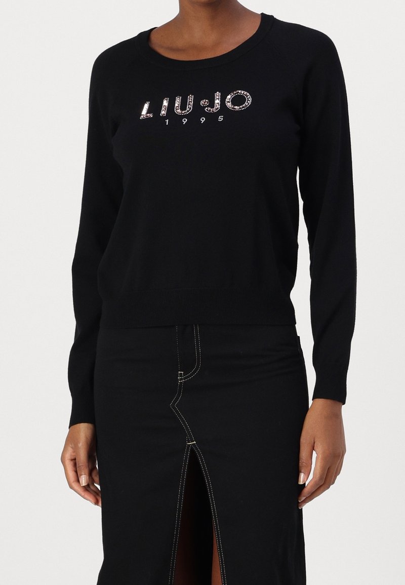 Femme portant un pull noir à manches longues avec l'inscription en strass « LIU JO 1995 » et une jupe noire avec des coutures blanches et une fente à l'avant.
