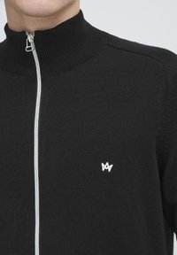 Maglione nero con zip, dotato di un colletto alto, con zip silver e piccolo logo bianco sul lato sinistro del petto. Tessuto in maglia morbida.