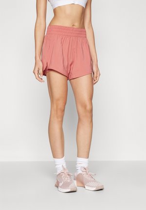 Mujer con pantalones cortos deportivos de color rosa claro, camiseta corta blanca, calcetines blancos y zapatillas de correr rosas, de pie contra un fondo liso.