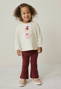 Lächelndes Kleinkind mit lockigem Haar, das einen cremefarbenen Pullover mit Ballerina-Design, bordeauxfarbene Hose und beige Turnschuhe auf neutralem Hintergrund trägt.