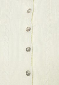 Gros plan sur un tissu tricoté crème avec des motifs de câbles verticaux et quatre boutons ronds transparents alignés en une seule rangée.