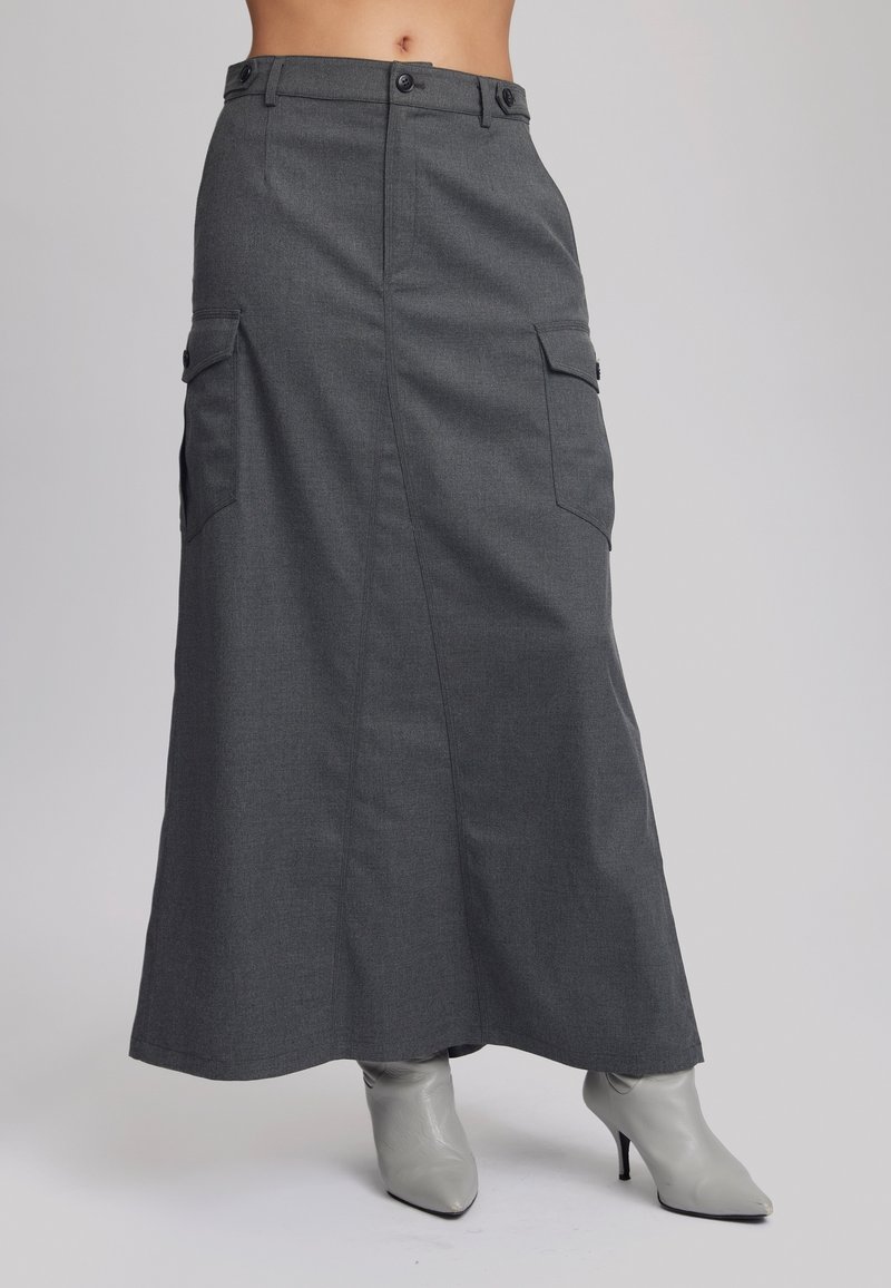 Jupe longue gris foncé avec poches latérales, portée avec des bottines à talons hauts pointues gris clair sur un fond uni.