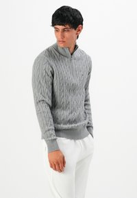 Mann in grauem, kabelgestricktem Halbzip-Pullover und weißen Hosen, der nach links unten schaut, vor einem schlichten weißen Hintergrund.