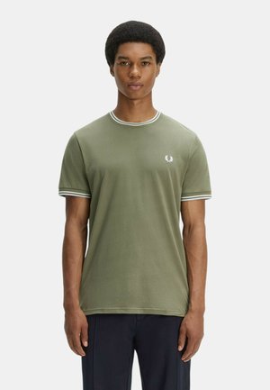 Junger Mann trägt ein olivgrünes T-Shirt mit weißer Umrandung am Kragen und an den Ärmeln, steht mit Blick nach vorne vor einem einfarbigen Hintergrund.