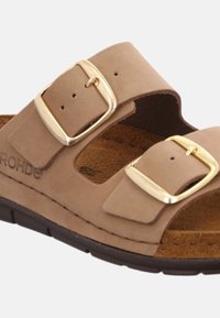 Brun mockaskinnsandalen med två justerbara guldförsedda spännen. Har en vadderad fotbädd och en texturerad gummisula.