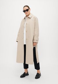 Beige trenčkot, zapínání na knoflíky, oversized střih, špičatý límec, boční rozparky; v kombinaci s černými kalhotami a mokasíny, černé sluneční brýle.