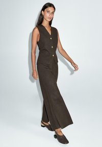 Veste marron sans manches avec boutons dorés et pantalon large assorti. Texture lisse, coupe ajustée, complétée par des chaussures décontractées.