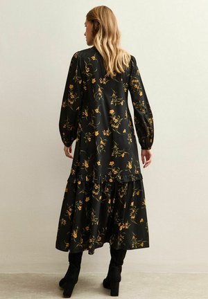 Vestito nero a fiori con maniche lunghe, orlo arricciato e silhouette fluida. Presenta motivi floreali gialli e verdi su uno sfondo scuro.