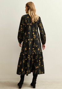 Zwarte bloemenjurk met lange mouwen, geruite zoom en een vloeiende silhouet. Kenmerkt zich door gele en groene bloemmotieven op een donkere achtergrond.