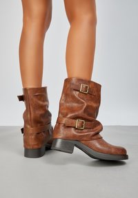 Bottines en cuir marron avec un design flou, dotées de boucles dorées et d'un talon bas. Surface texturée avec une finition mate.