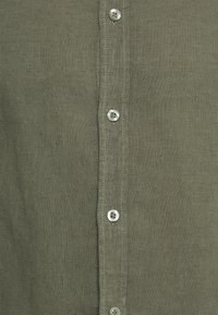 Camicia di lino verde oliva con abbottonatura a lunghezza intera, bottoni in tonalità argento e cuciture visibili lungo i bordi.