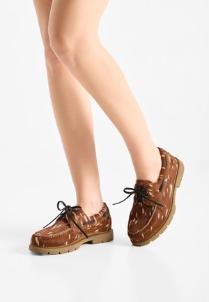 Gambe che indossano scarpe stile mocassino marrone con macchie bianche, lacci neri e suole spesse color beige su uno sfondo bianco semplice.