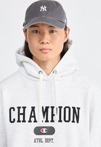 Sivá šiltovka s bielym logom, biela mikina s čiernym textom a logom "CHAMPION". Materiál vyzerá mäkko, má šnúrky a voľný strih.