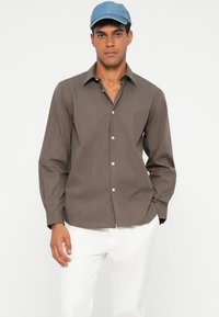 Camisa de botones a rayas marrón con cuello, mangas largas y botones blancos, combinada con pantalones blancos y una gorra azul.