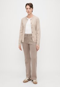 Bēšas krāsas kardigāns ar pogām, uzvilkts virs baltas T-krekla, kombinēts ar gaiši bēšām biksēm un bēšiem sporta apaviem, uz vienkārša fona.