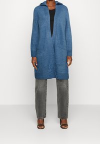 Lång, blå stickad cardigan med huva, med ett texturerat mönster, två framfickor och ribbade mudd. Parat med mörkgrå jeans och klackar.