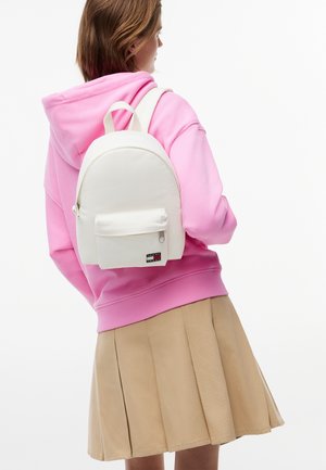 Persona con sudadera con capucha rosa y falda plisada beige, llevando una pequeña mochila blanca con un bolsillo delantero y logotipo.
