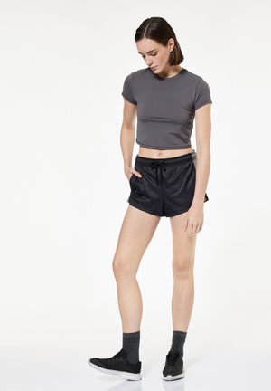 Młoda kobieta w szarym krótkim topie, czarnych sportowych spodenkach, ciemnych skarpetkach i czarnych sportowych butach, stoi z jedną ręką w kieszeni, głowa opuszczona.