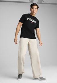 Sort bomulds T-shirt med "PORTUGAL" tekst og rød logo, parret med lyse beige brede bukser og grå sneakers. Enkel, afslappet outfit.