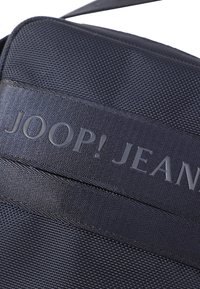 Temno moder tekstilni messenger nahrbtnik z teksturirano tkanino, ki se ponaša s poudarjenim odtisnjenim logotipom "JOOP! JEANS" na gladkem traku. Vključena nastavljiva naramnica.
