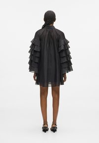 Μαύρο διάφανο φόρεμα με πτυχωτά ρυάκια στους ώμους, ψηλό λαιμό και χαλαρή εφαρμογή. Το υλικό φαίνεται ελαφρύ και ημιδιαφανές.