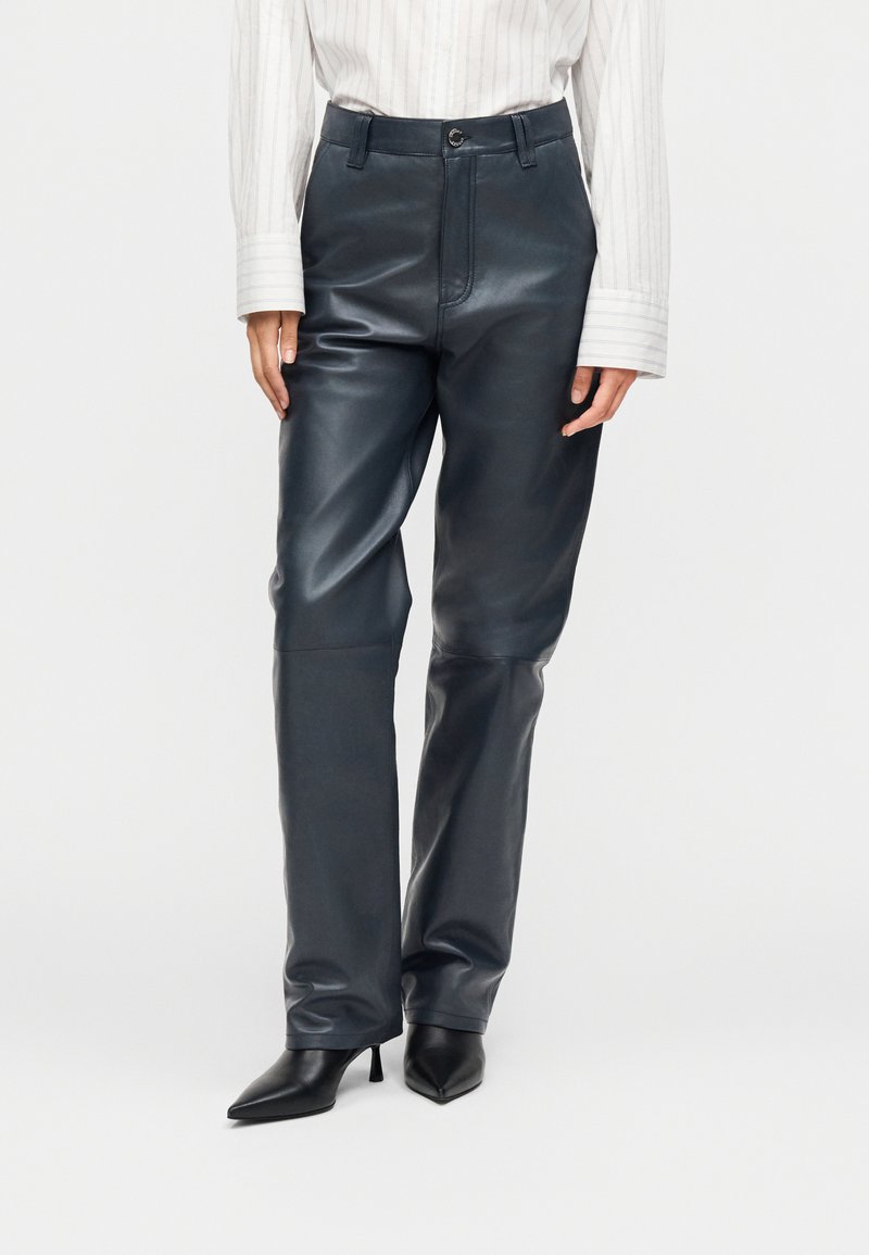 Femme portant un pantalon en cuir gris foncé à taille haute, un chemisier blanc à manches longues et des bottes noires à talons pointues, debout devant un fond blanc.