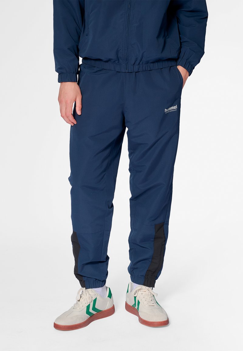 Mörkblå träningsbyxor i lätt material, med elastiska muddar, sidfickor och svarta detaljer längs benen. Synligt märkeslogotyp.