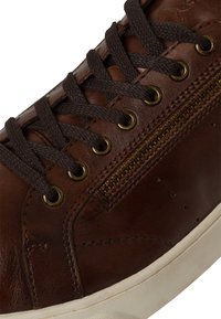 Baskets en cuir marron avec des lacets marron foncé, des œillets en laiton et une fermeture éclair sur le côté. La semelle est blanche et légèrement texturée.