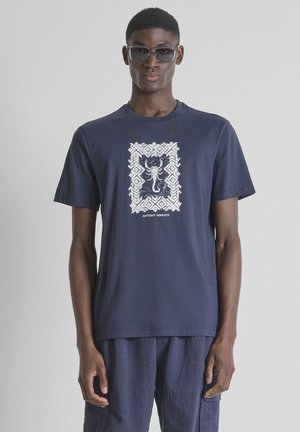 Hombre con camiseta azul marino con un diseño de escorpión blanco enmarcado por un patrón geométrico, pantalones texturizados en azul marino y gafas de sol oscuras.