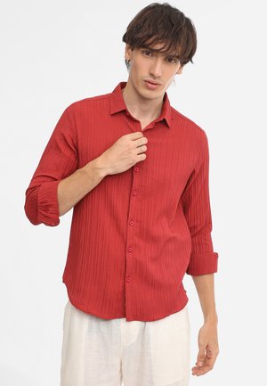 Rot gestreifte Bluse mit langen Ärmeln, die an den Ellbogen aufgerollt sind, Knopfleiste mit rosa Knöpfen, aus leichtem Stoff. Darunter weiße Hose.