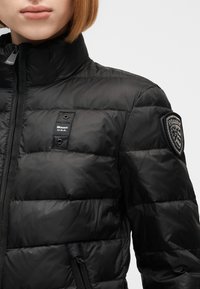 Femme portant une veste matelassée noire avec un col montant, une fermeture éclair à l'avant, un logo Blauer U.S.A. sur la poitrine et un écusson sur la manche.