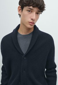 Navy ribbed cardigan with a shawl collar, featuring five buttons, worn over a gray crewneck sweater, highlighting texture and layering.