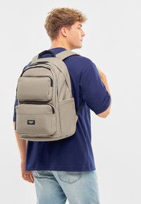 Fiatal férfi kék pólóban és világos farmernadrágban, bézs hátizsákkal, amelynek több cipzáros rekesz van, jobbról hátról nézve.