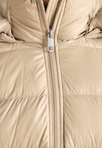 Beige vadderad jacka fram med silverfärgad dragkedja och fäst fuskpäls vid kragekanterna.