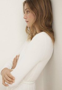 Robe blanche à manches longues avec une texture lisse, une silhouette ajustée et une taille bien marquée. Les bras du modèle sont croisés contre un mur.