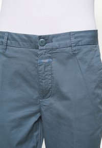 Pantalones de algodón azules con frente plano, cierre de botón y una etiqueta en la cintura, con textura suave y un brillo sutil.