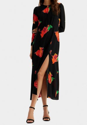 Femme portant une robe maxi noire à manches longues avec des imprimés floraux rouges et verts et une fente haute sur la cuisse, associée à des talons à bride cheville noirs.