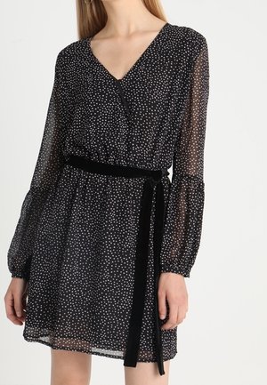 Mujer con un vestido negro con lunares blancos, mangas largas transparentes y un cinturón negro en la cintura, de pie contra un fondo claro y sencillo.