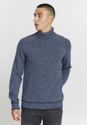 Navy blauwe gebreide trui met hoge kraag, geribde manchetten en zoom; textiel met een subtiel patroon; gedragen over donkergrijze broek.