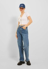 Top corto bianco a maniche corte con bottoni sul davanti, abbinato a jeans denim blu larghi e scarpe platform nere chunky, completato da un cappellino blu.