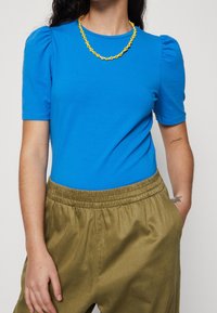 Blå kortärmad topp med puffade axlar, kombinerad med löst sittande olivgröna byxor. Outfiten pryds av ett gult blommigt halsband.